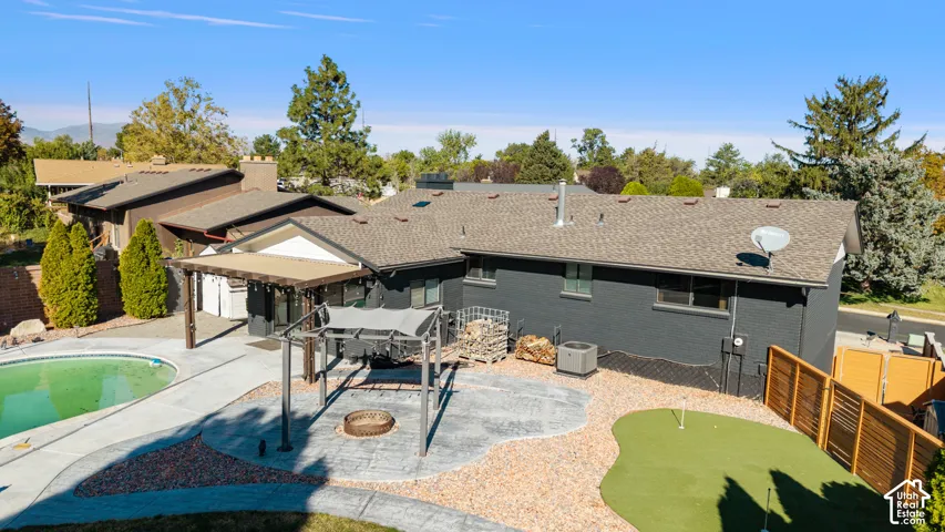 Expansive backyard oasis featuring a resort-style swimming pool with integrated spa, manicured putting green, multi-level paver patio with pergola cover, mature landscaping with evergreen trees, and a modern single-story home with dark exterior finishes a