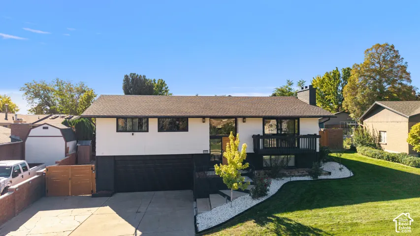 Contemporary split-level home featuring a two-car garage with dark finish, crisp white and charcoal exterior, expansive manicured lawn with mature landscaping, and a composite deck with black railings overlooking the grounds