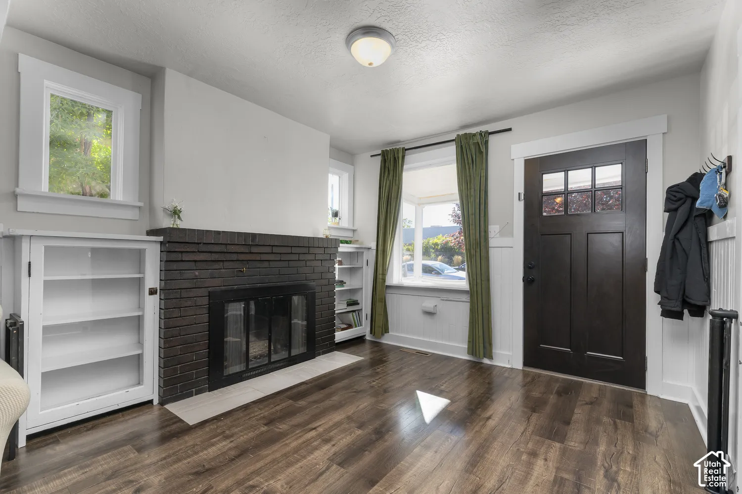 Dark wood-finish flooring, brick fireplace with tile hearth, built-in shelving, white beadboard wainscoting, and a dark paneled entry door with glass inserts
