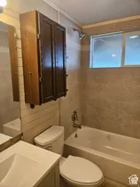 White porcelain bathtub with chrome fixtures, tiled shower surround, dual-pane window, and a built-in wooden cabinet