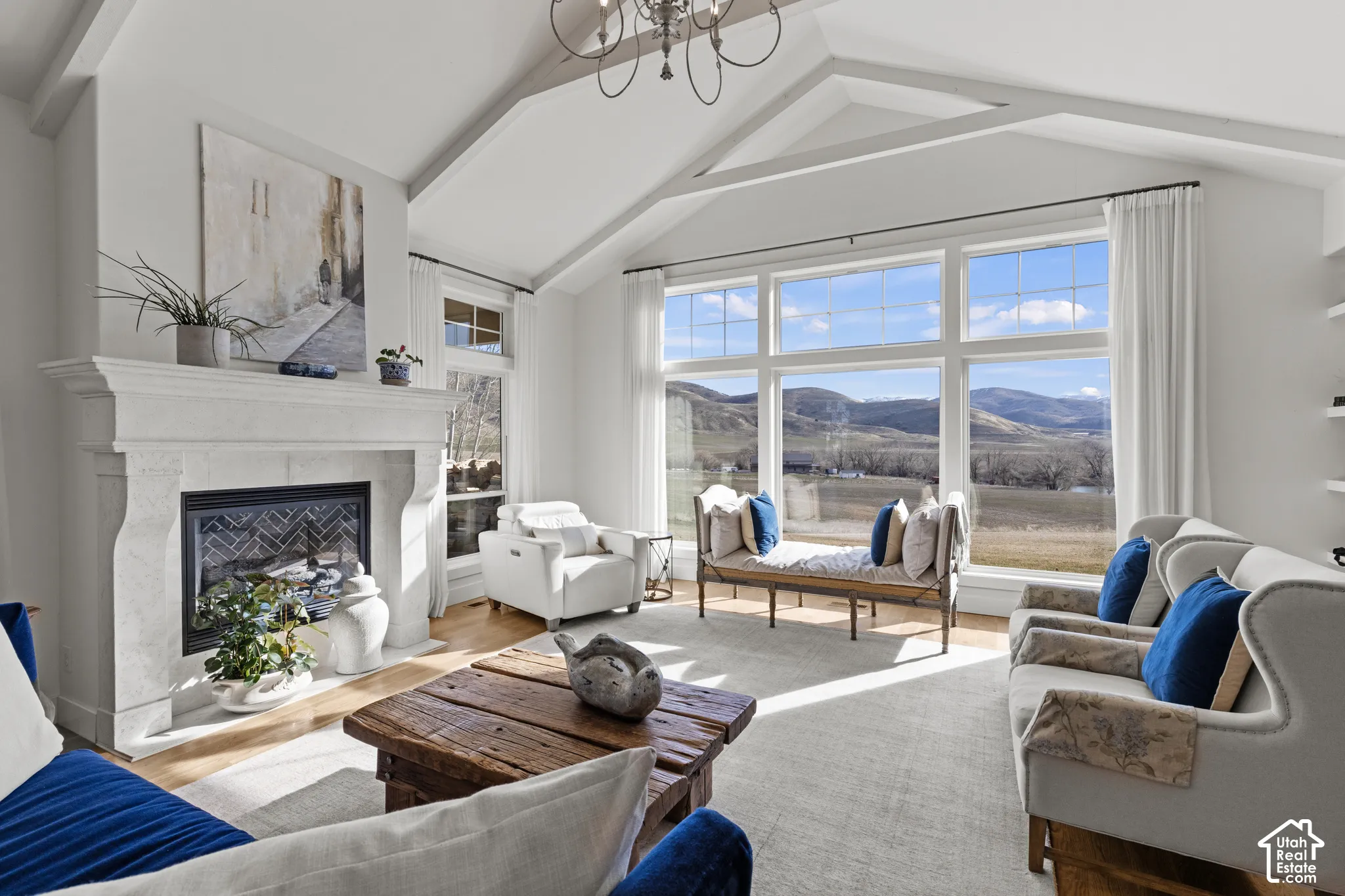 Bright, open living room featuring vaulted ceilings with exposed beams, a white brick fireplace with wood-burning insert, and expansive multi-pane windows framing mountain and valley scenery