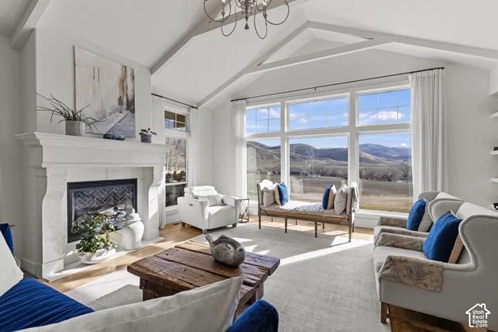 Bright, open living room featuring vaulted ceilings with exposed beams, a white brick fireplace with wood-burning insert, and expansive multi-pane windows framing mountain and valley scenery
