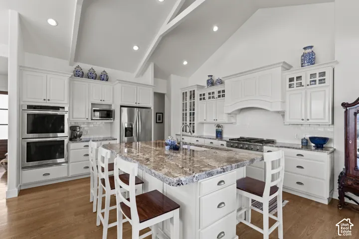 Vaulted ceiling with exposed beams, white cabinetry, integrated double ovens, large kitchen island with counter seating, and wood-finish flooring