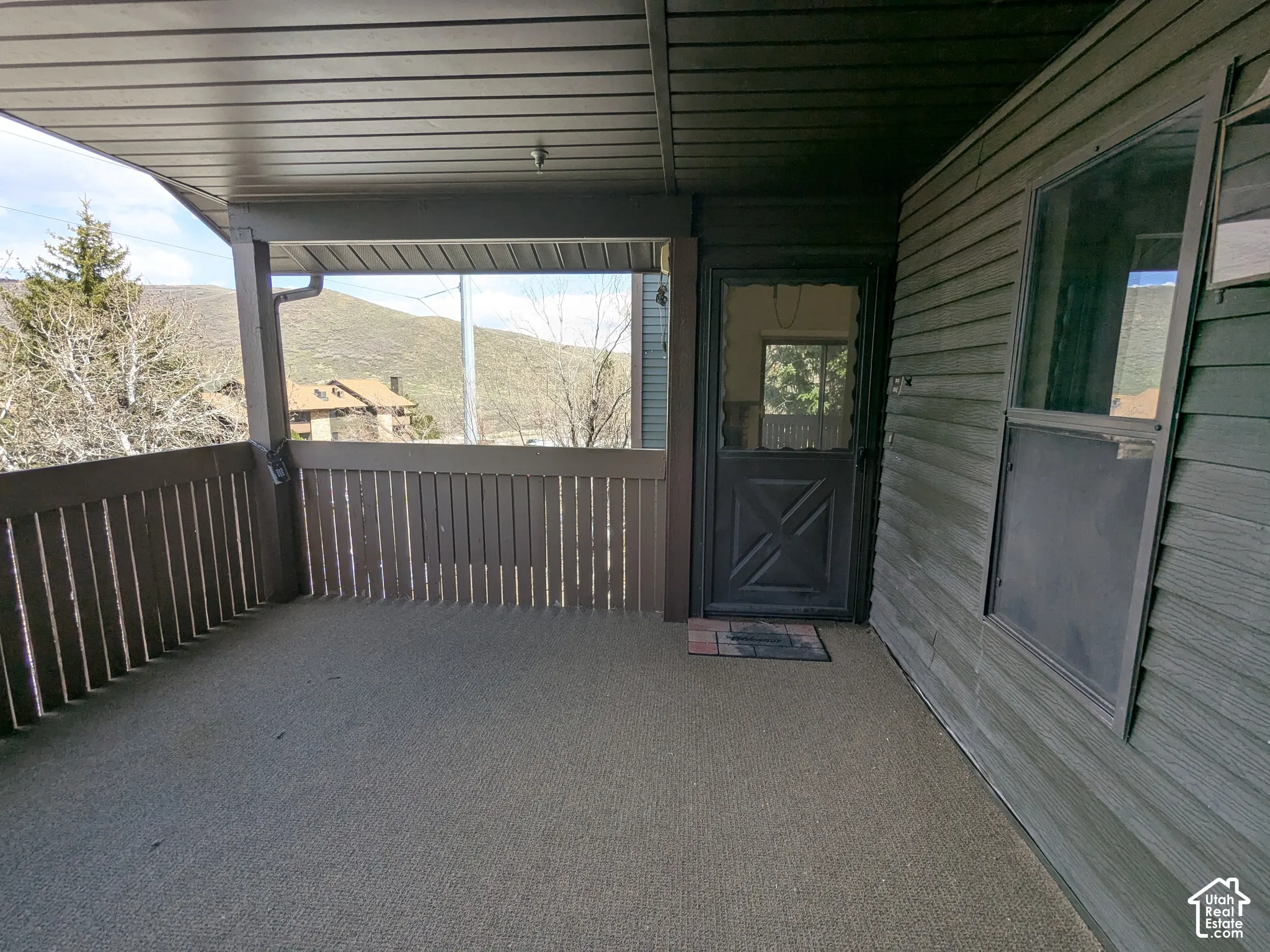 Covered outdoor patio with slatted railing, exterior siding, and a solid entry door with an upper glass pane