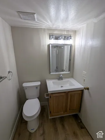 Wood-finish flooring, single vanity with a white countertop and natural wood cabinetry, polished chrome faucet, rectangular mirror, and a polished chrome light fixture
