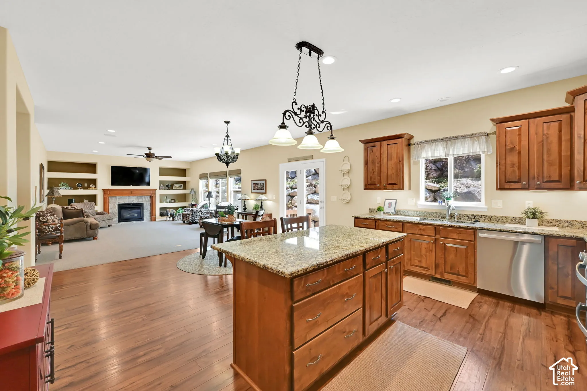 Wood-finish flooring, central kitchen island with a light-toned countertop, wood cabinetry, stainless steel appliances, and a fireplace with a mantel