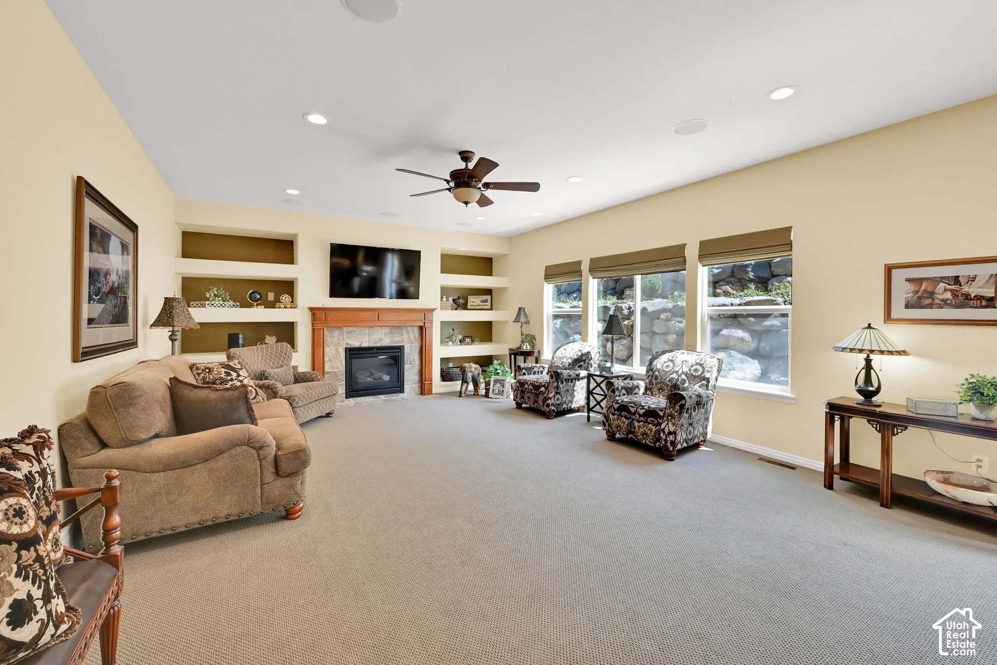 Spacious room featuring a central fireplace with a wood mantel and stone surround, built-in shelving, recessed lighting, and multiple windows