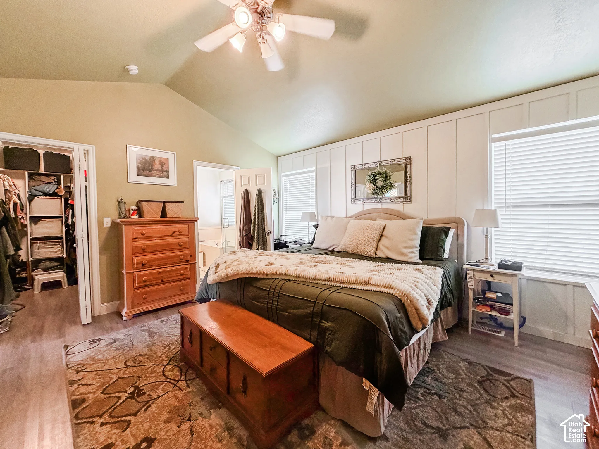 Vaulted ceiling with ceiling fan, wood-finish flooring, accent wall with wainscoting, two windows with blinds, and an open closet