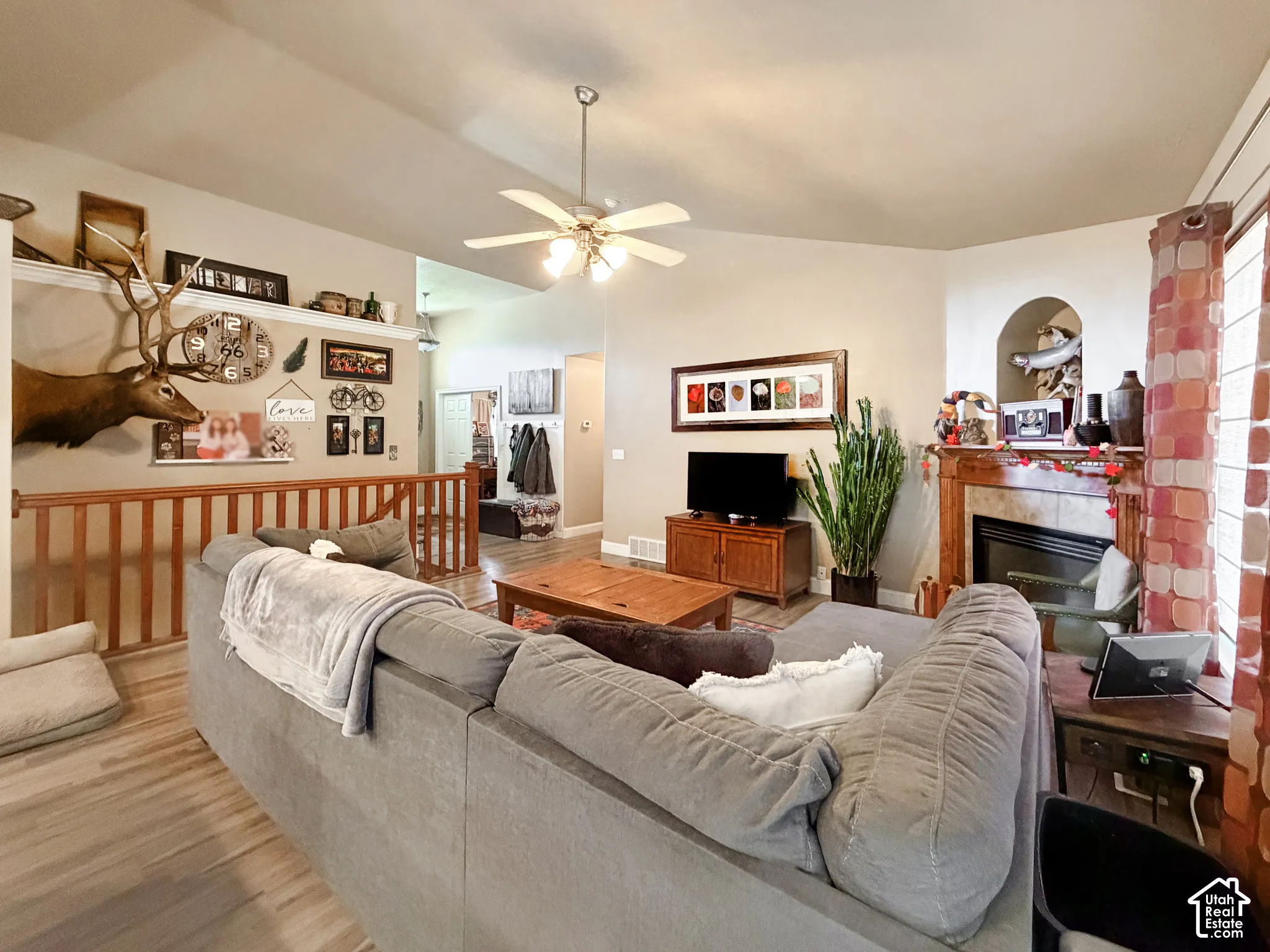 Vaulted ceiling with ceiling fan, wood-finish flooring, built-in fireplace, staircase with wood railing, and window with curtains