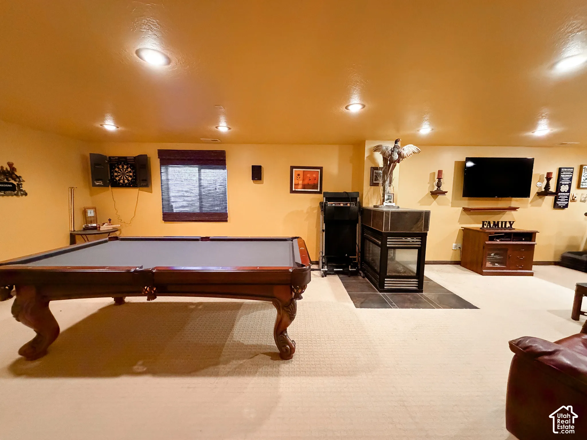 Recessed lighting illuminates the room, featuring a built-in fireplace with a stone surround, a mounted television, a window with blinds, and carpeted flooring