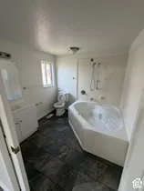 Corner soaking tub, dark tile flooring, white vanity with integrated sink, wall-mounted mirror, and toilet