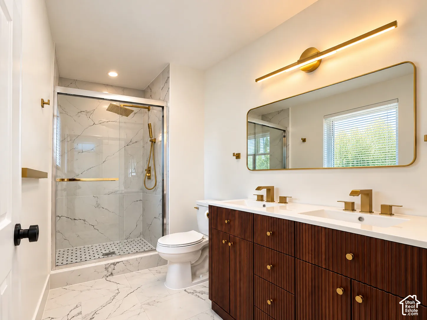 Dual vanity with gold-finish hardware and integrated sinks, walk-in shower with glass enclosure and marble-look tile, gold-finish linear sconce, and gold-frame mirror