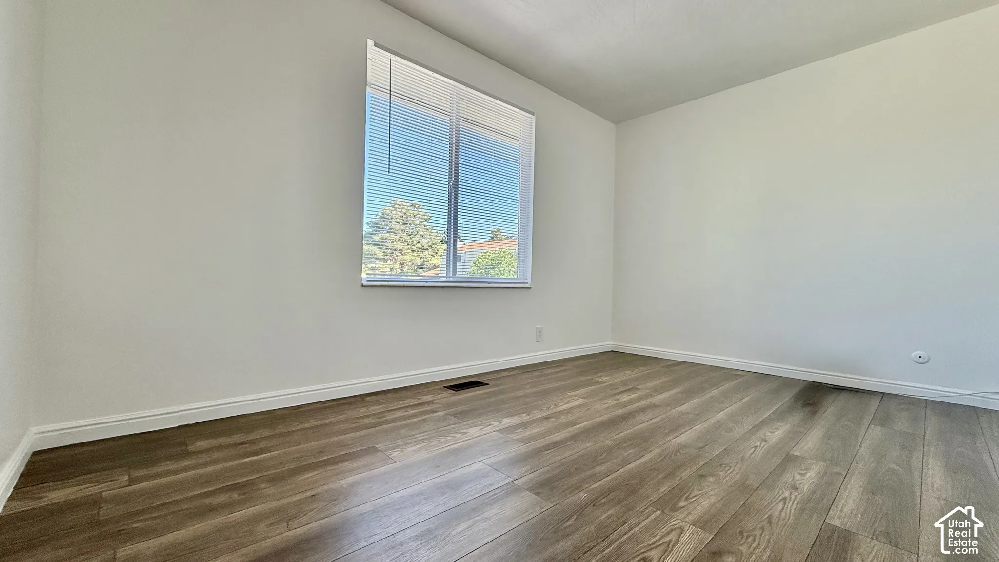 Spacious room featuring wood-finish flooring, white walls, and a large window with horizontal blinds