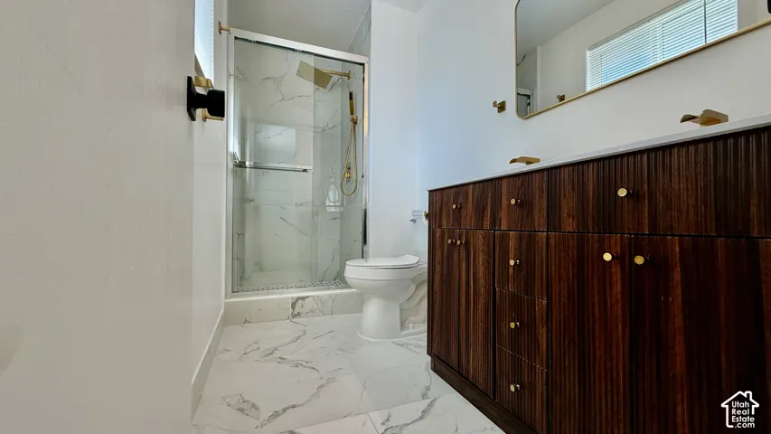 White and grey patterned floor tiling, glass-enclosed shower with gold-finish fixtures, white toilet, dark wood-finish vanity with fluted detailing, gold-finish hardware, and a framed mirror