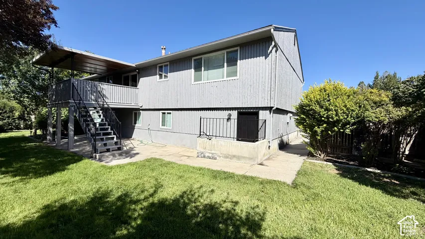 Rear exterior featuring gray siding, a covered upper-level deck, exterior stairs, a ground-level patio, and surrounding landscaping