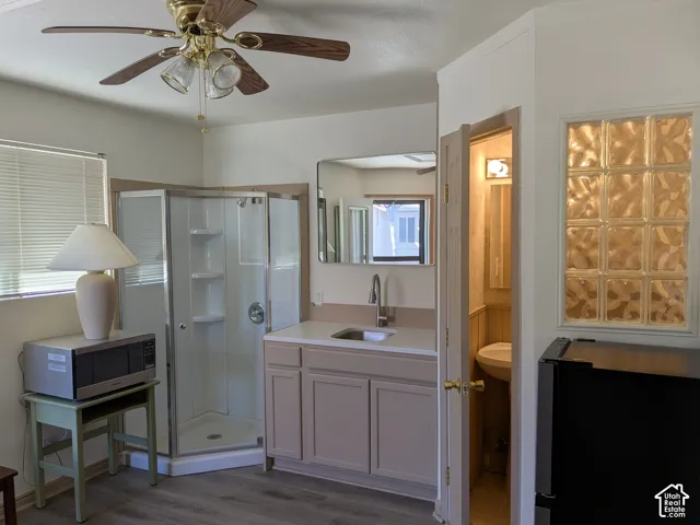 Wood-finish flooring, corner shower enclosure, vanity with integrated sink and chrome faucet, glass block window, and ceiling fan