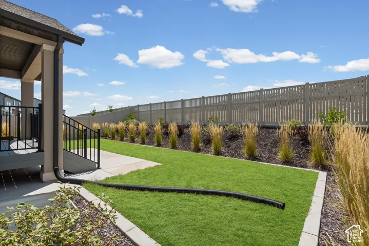 Covered patio with black railing, concrete patio, expansive lawn, garden beds with ornamental grasses, and perimeter fencing