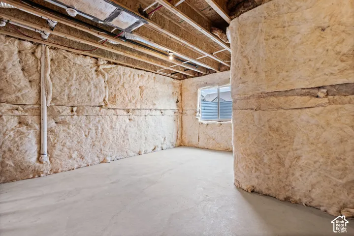 Unfinished basement featuring exposed wood joists, batt insulation, a concrete floor, and a window with a well