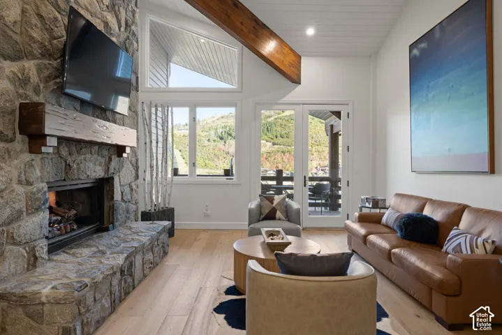 Stone-clad fireplace with a substantial timber mantel, vaulted ceiling with exposed timber beam, wood-finish flooring, and white French doors leading to an outdoor space