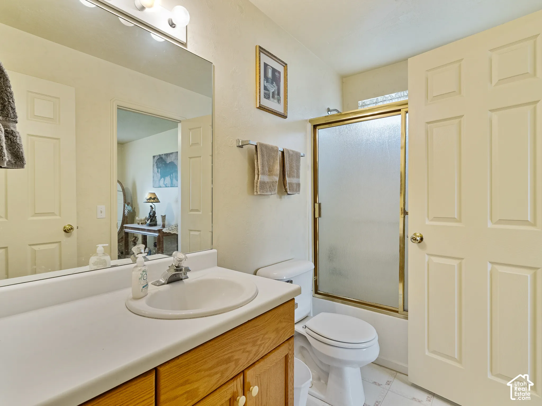 Bathroom featuring a wood-finish vanity with a white countertop and integrated sink, a large wall-mounted mirror, a three-bulb vanity light, and a shower with a gold-finish enclosure