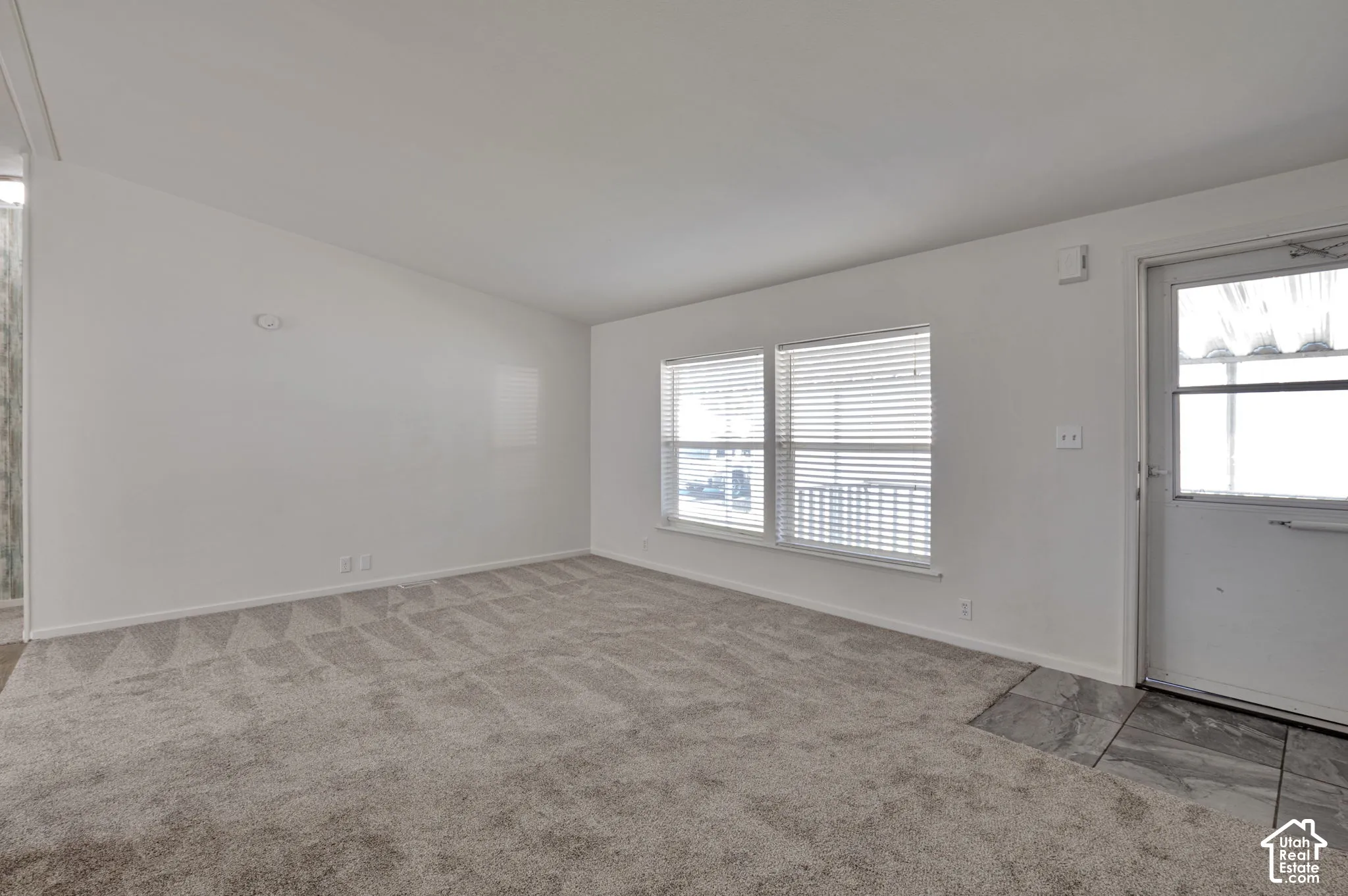 Spacious room with white walls, patterned carpet flooring, dual windows with blinds, and an entry door