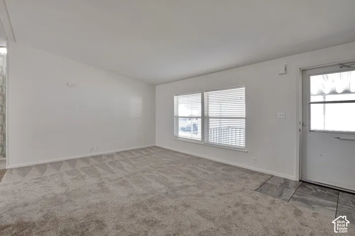 Spacious room with white walls, patterned carpet flooring, dual windows with blinds, and an entry door