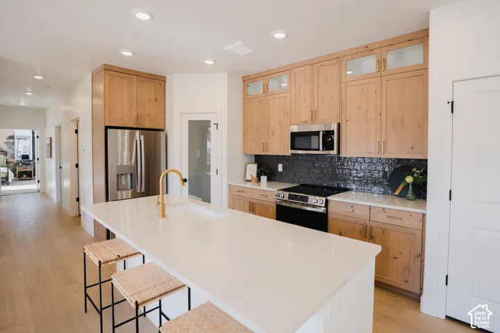 Kitchen featuring a white countertop island, integrated sink with a gold-finish faucet, light wood-finish cabinetry, and a dark tile backsplash