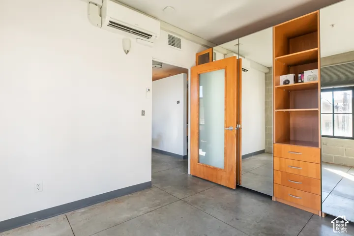 Concrete flooring, wood-finish door with frosted glass panel, integrated shelving unit with drawers, mirrored wall, and wall-mounted air conditioning unit