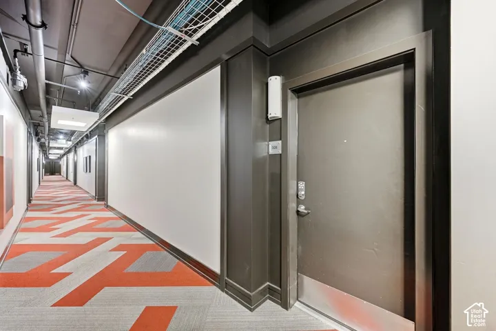 Corridor featuring patterned carpet, white wall panels, and exposed ceiling utilities
