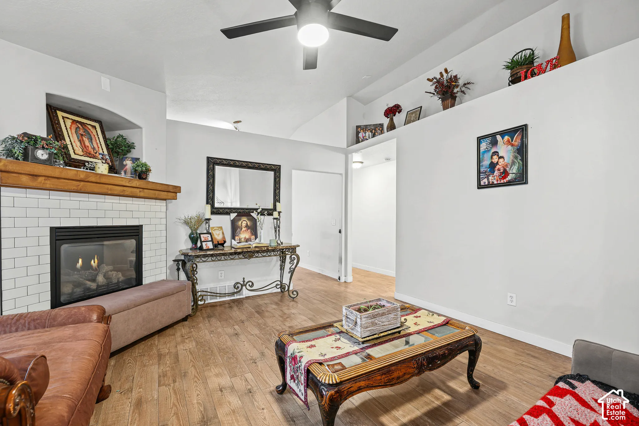 Wood-finish flooring, vaulted ceiling, built-in fireplace with subway tile surround, recessed display shelf, and ceiling fan with integrated lighting