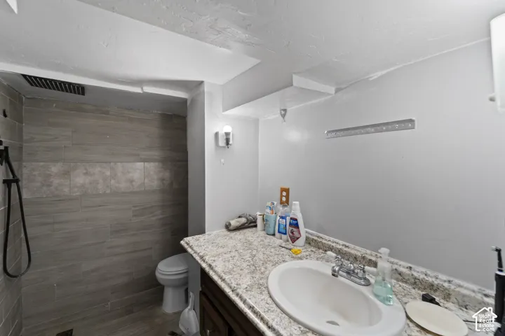 Walk-in shower with wood-finish tile, toilet, vanity with integrated sink, and wall-mounted lighting