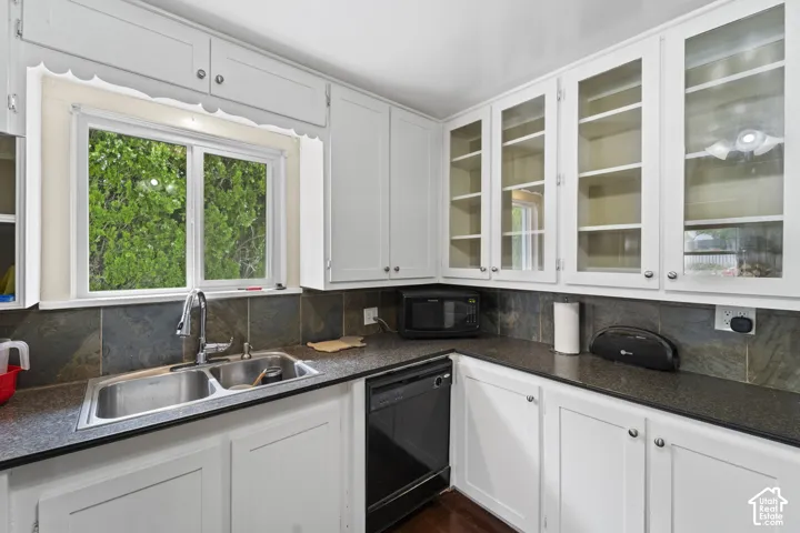 White cabinetry with glass-front uppers, stainless steel double basin sink, dark stone backsplash, and dark countertops