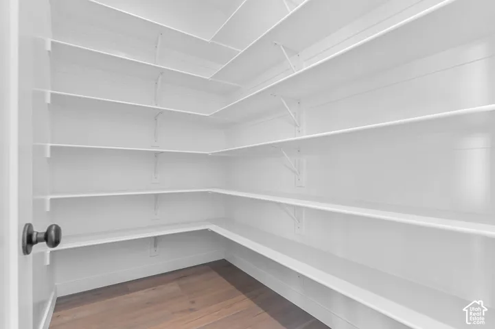 Walk-in pantry featuring extensive white shelving, dark wood-finish flooring, and a matte black door handle