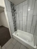 Built-in bathtub with shower, white subway tile surround, gray mosaic accent tile, and tiled flooring