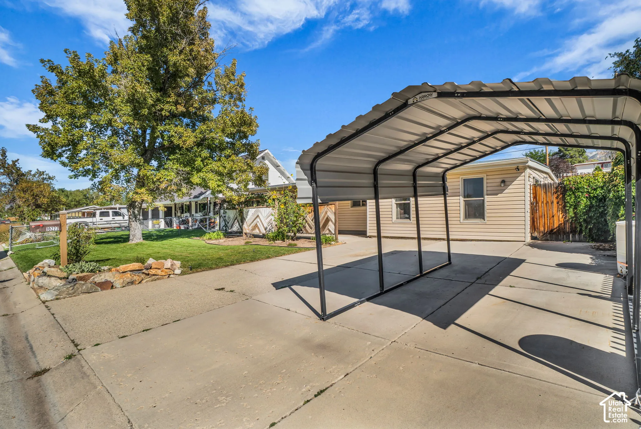 Carport structure, concrete driveway, mature trees, and natural stone landscaping elements