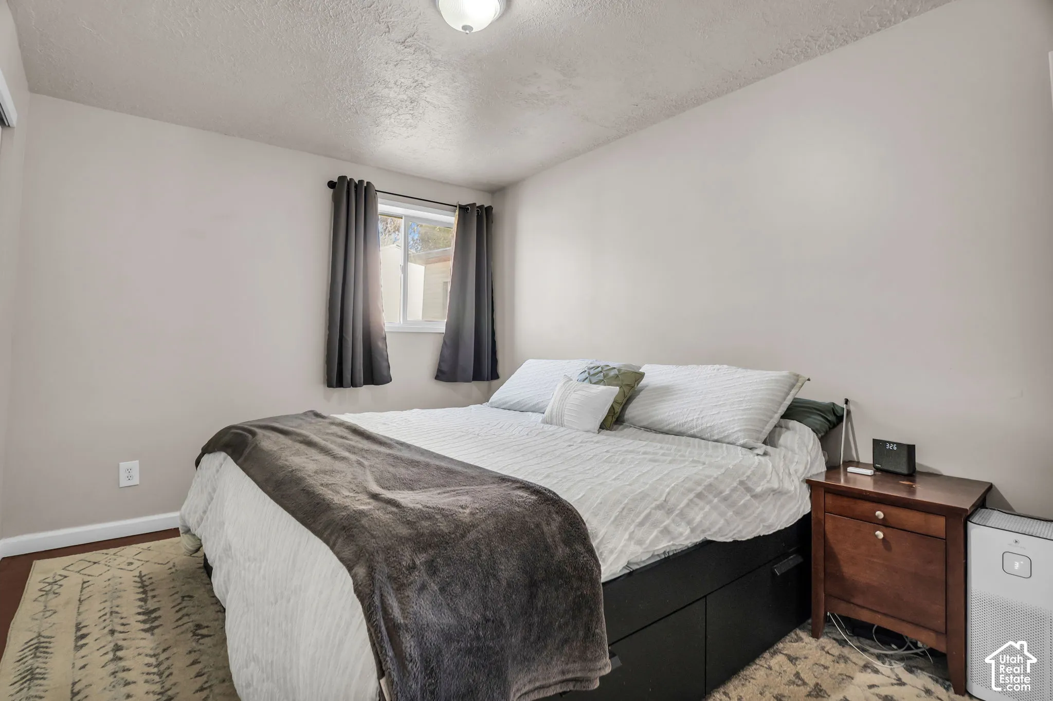 Bright secondary bedroom featuring a large window with blackout curtains, neutral wall finishes, hardwood-finish flooring accented with area rugs, and wood nightstand with drawer storage