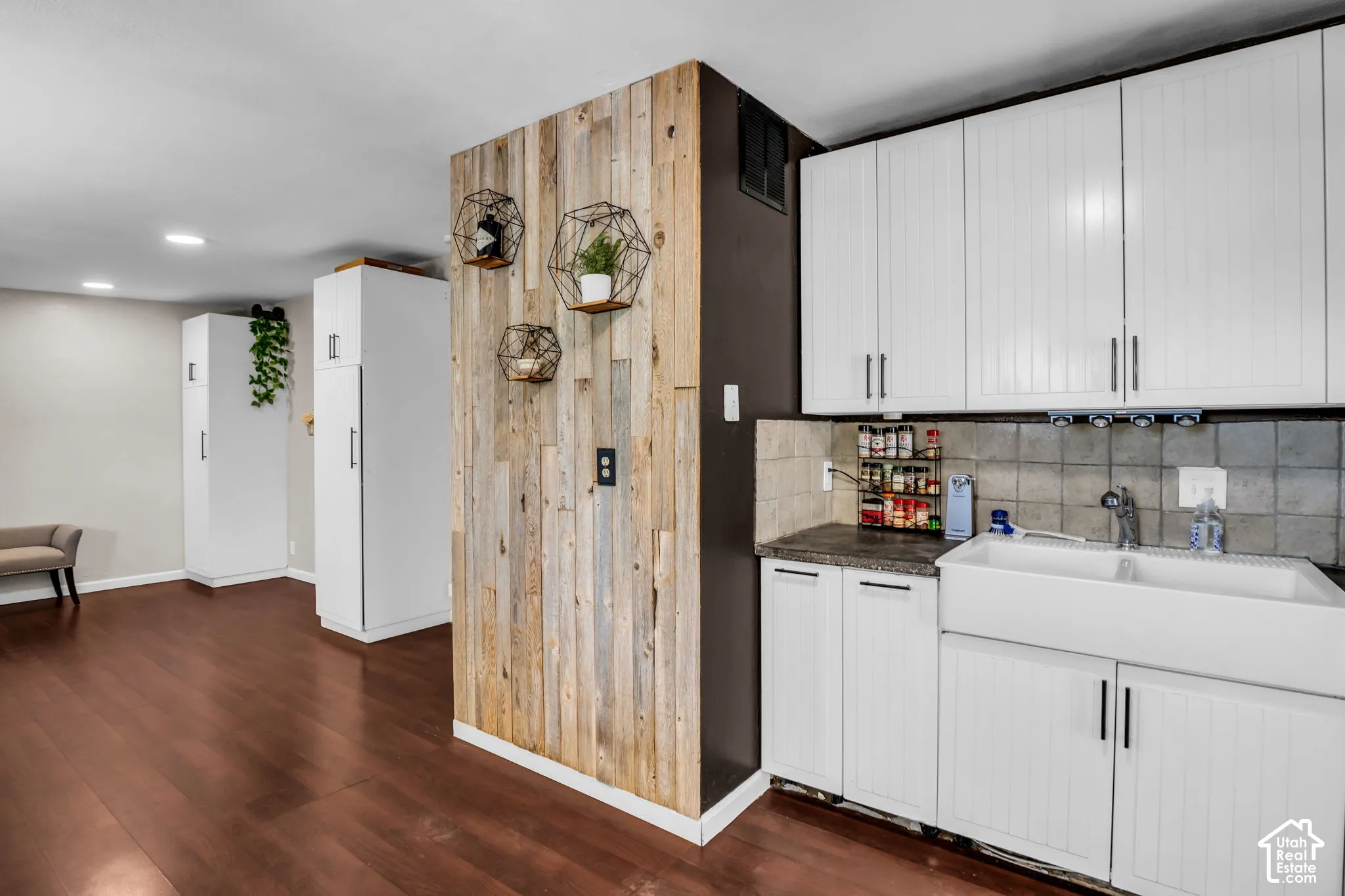 Wood-finish flooring, white cabinetry, farmhouse sink, light gray tile backsplash, and a wood-paneled accent wall