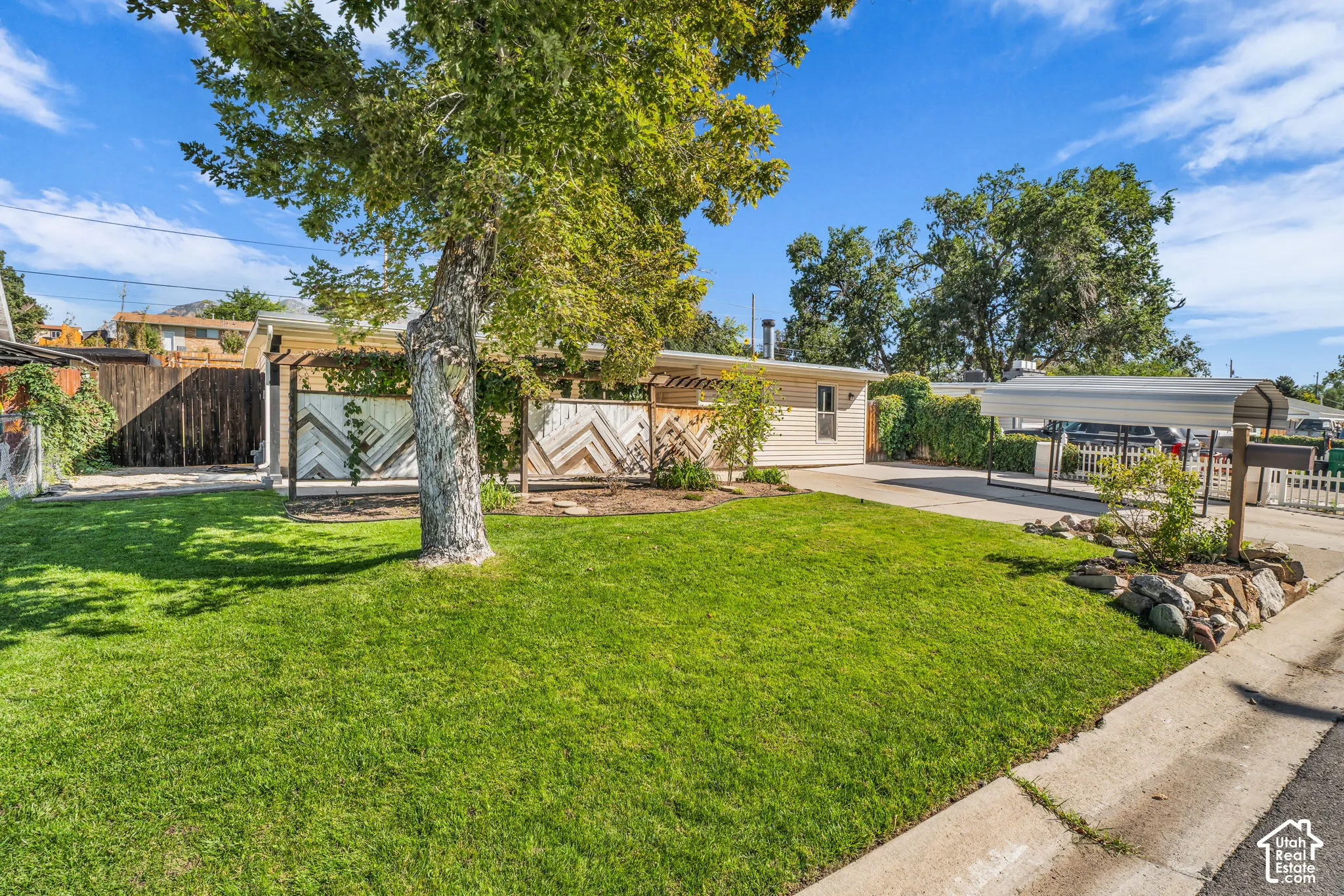 Manicured lawn, mature trees, decorative privacy fencing, extended driveway, and carport