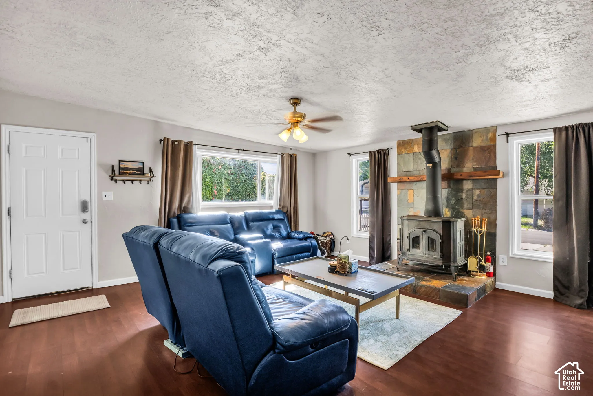 Wood-finish flooring, built-in wood-burning stove with stone surround, ceiling fan, and multiple windows