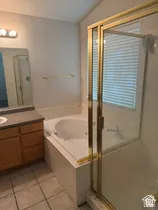 Tiled flooring, built-in jetted tub, wood-finish vanity with countertop, and an enclosed shower with gold-tone framing