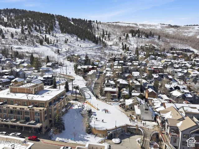 Snow-covered mountain landscape with dense evergreen tree coverage, diverse residential architecture, multistory commercial buildings, and a prominent pedestrian bridge