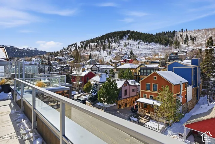Rooftop deck featuring glass railings and stone flooring, overlooking a snow-covered mountain landscape and a neighborhood with diverse architectural styles