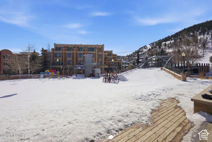 Snow-covered ground with a wood-plank pathway, a ski lift structure, a pedestrian bridge, and multi-story buildings with brick and stone facades