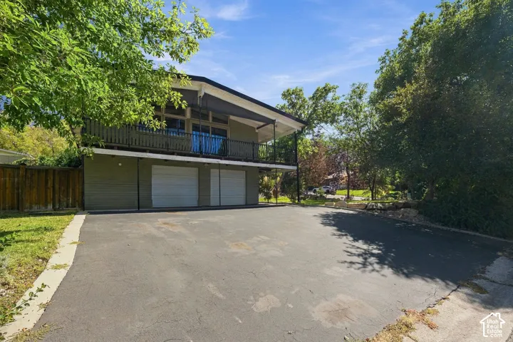 Two-story exterior with a covered upper balcony, ground-level two-car garage, expansive paved driveway, and established surrounding trees
