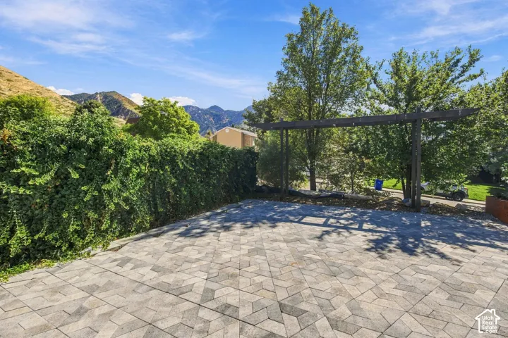 Paved patio area with a geometric paver design, mature perimeter landscaping, and a pergola structure