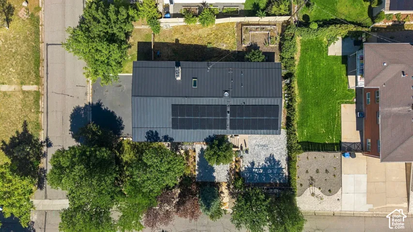 Modern metal roof with integrated solar panels, asphalt driveway, and a front yard featuring mature trees and a stone patio