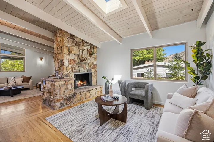 Stone-clad fireplace with hearth, wood-finish flooring, exposed beam ceiling, skylight, and large window