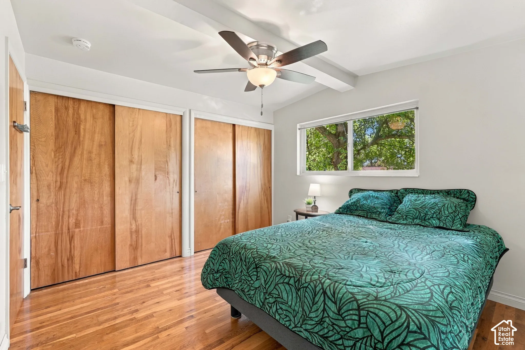 Vaulted ceiling with exposed beam, wood-finish flooring, dual sliding closet doors, ceiling fan with integrated lighting, and a horizontal window
