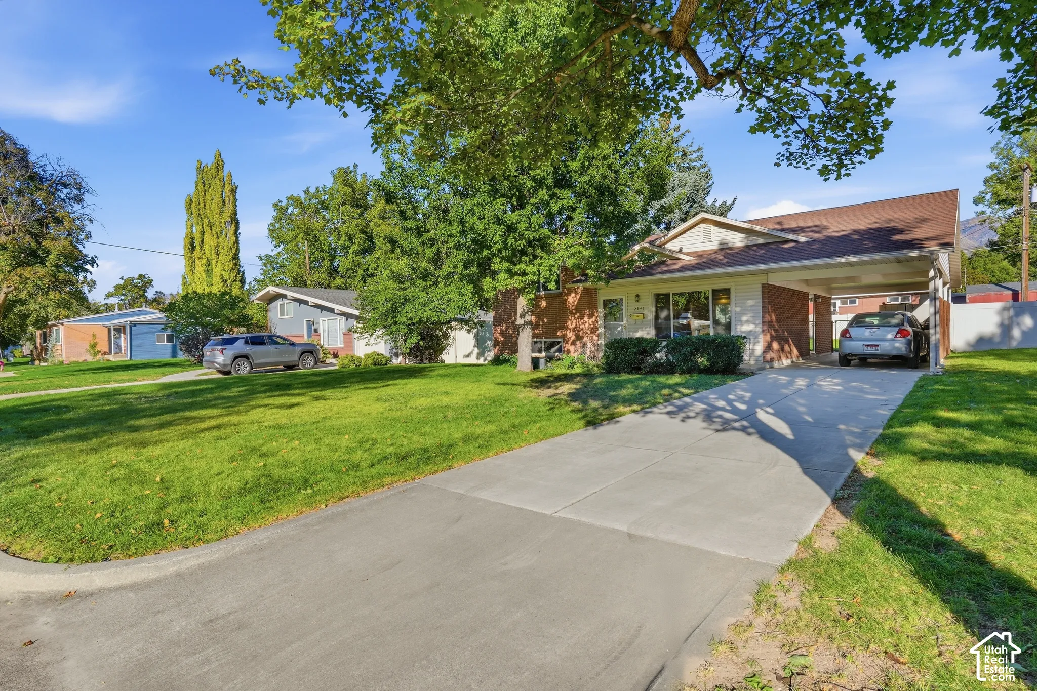 Brick and siding exterior with a covered carport, mature trees, and a concrete driveway