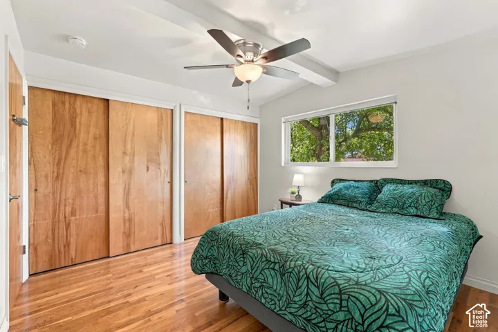 Vaulted ceiling with exposed beam, wood-finish flooring, dual sliding closet doors, ceiling fan with integrated lighting, and a horizontal window
